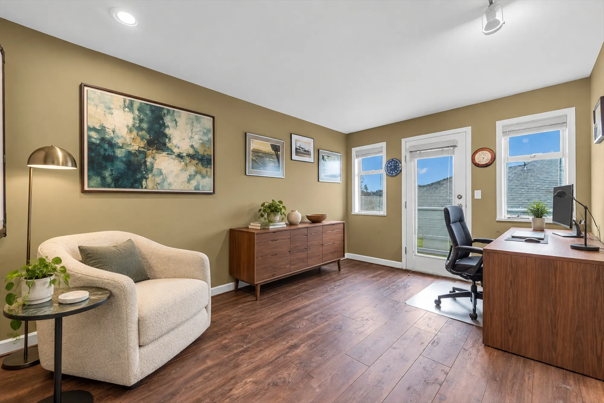 The same bedroom virtually staged as an office: an armchair and side table, a sideboard, a desk and chair, a floor lamp, a large painting and small framed pictures