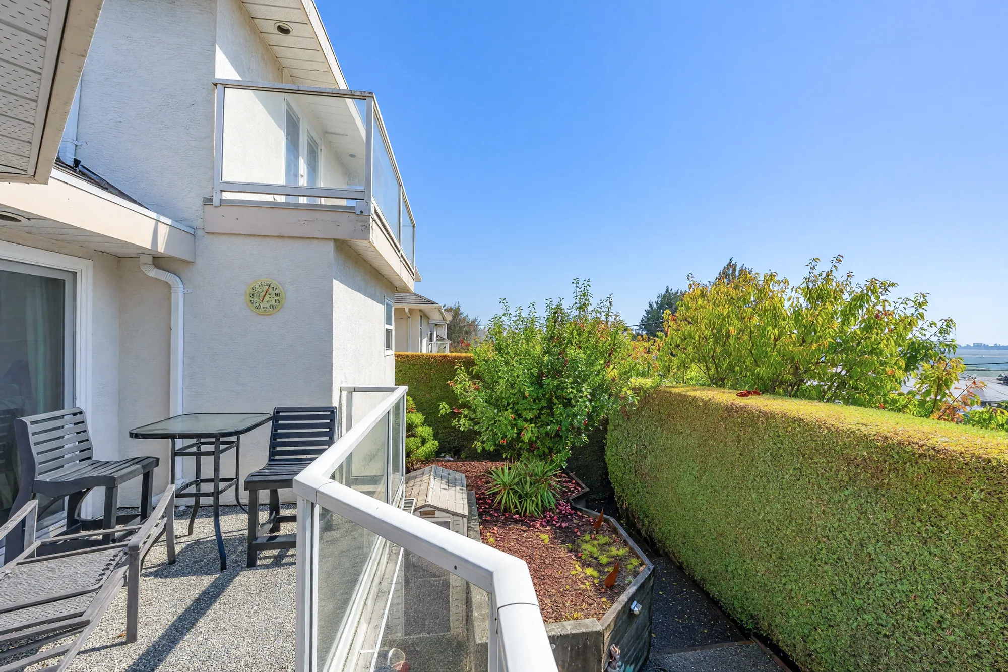 The deck beside a sliding door under the upstairs balcony: a glass table and chairs, glass railings, a hedge and a fruit tree