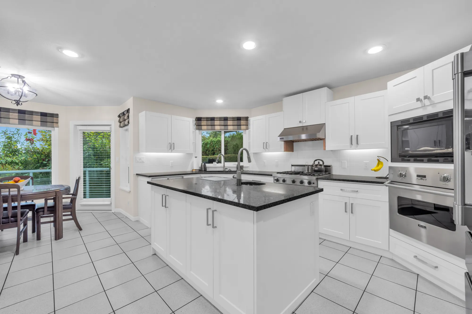 The kitchen: white shaker cabinets, dark grey counters, an island, a stainless gas range under a hood, a built-in oven and microwave, a tiled floor
