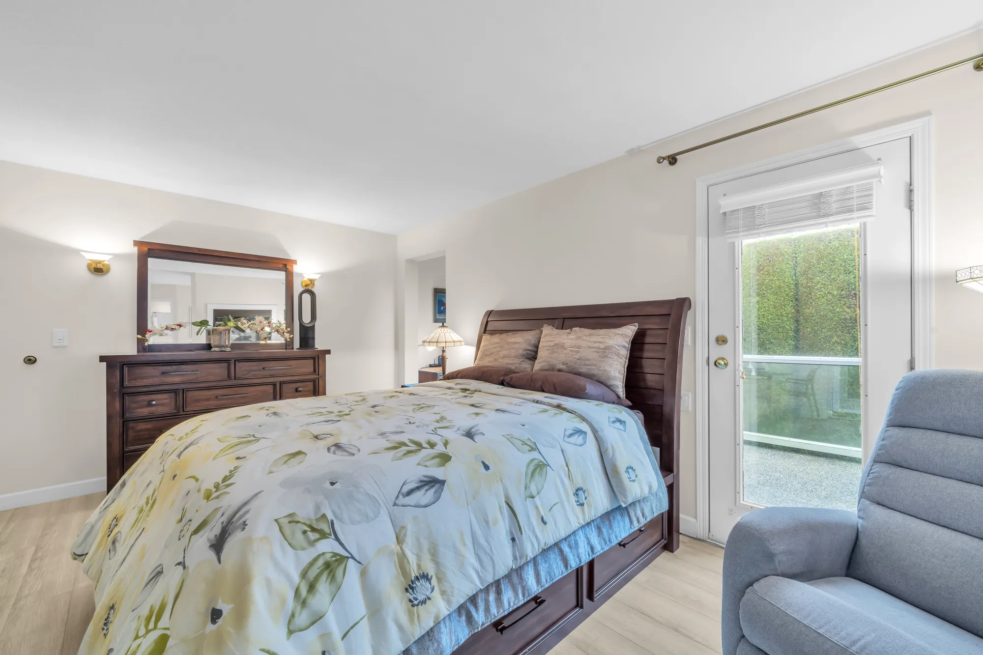 The primary bedroom: a bed with a floral cover, a dresser and mirror, a glass door to a balcony, a recliner