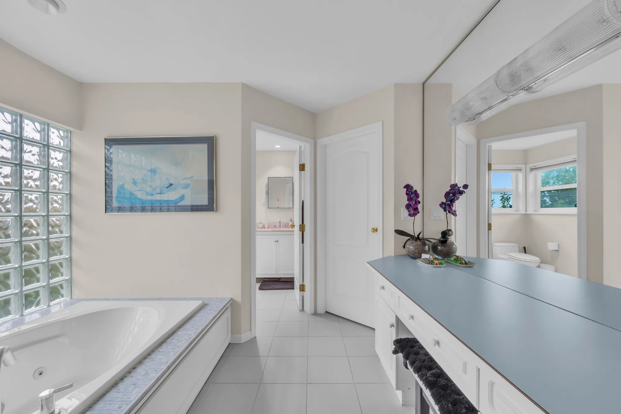 The primary bathroom: a jetted tub beside a glass-block window, a long vanity with a blue-grey counter, a tiled floor