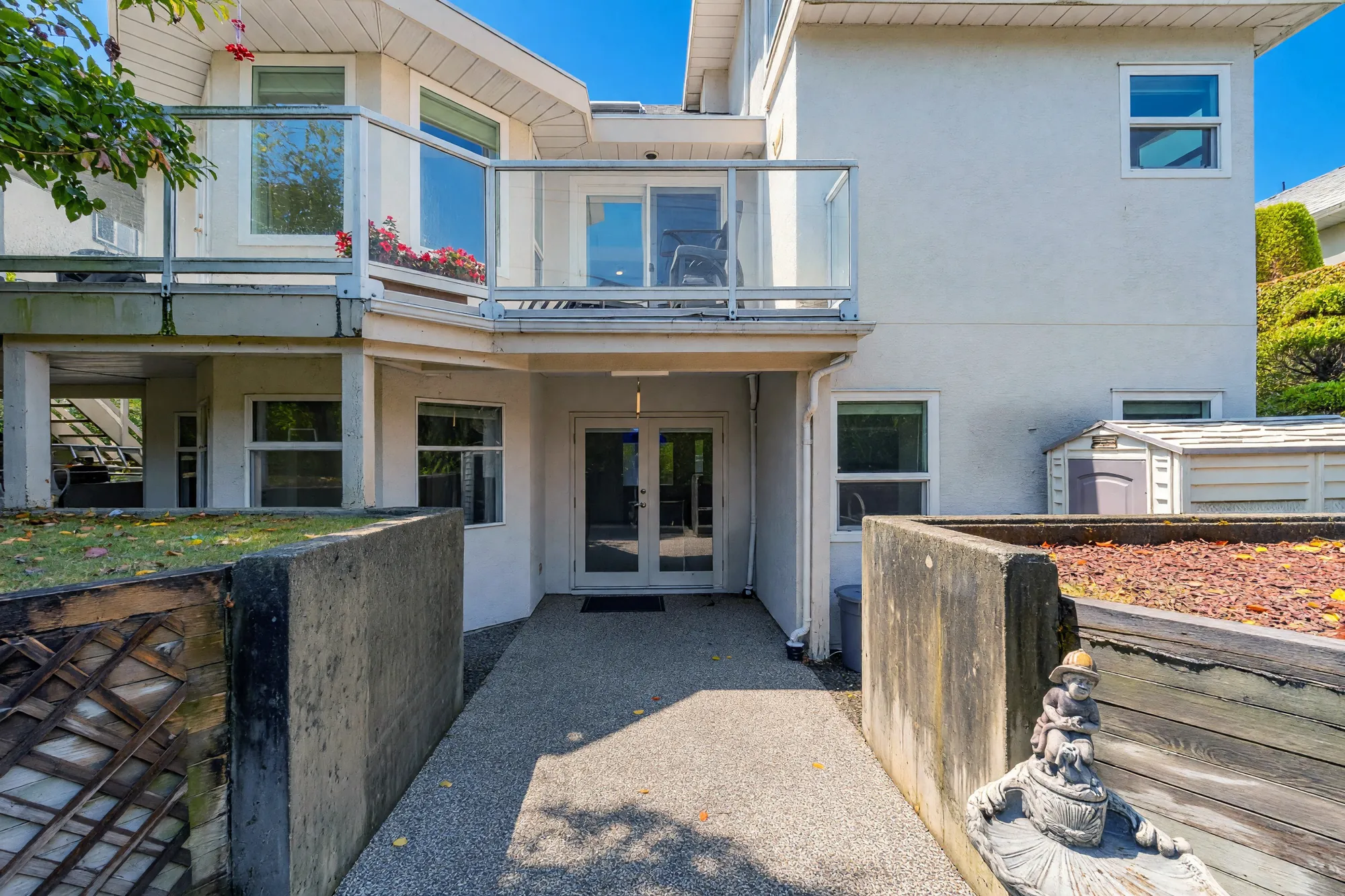 The back of the house at the lower floor: a pair of glass doors under the balcony, a paved path between raised planters, a small storage shed at the right